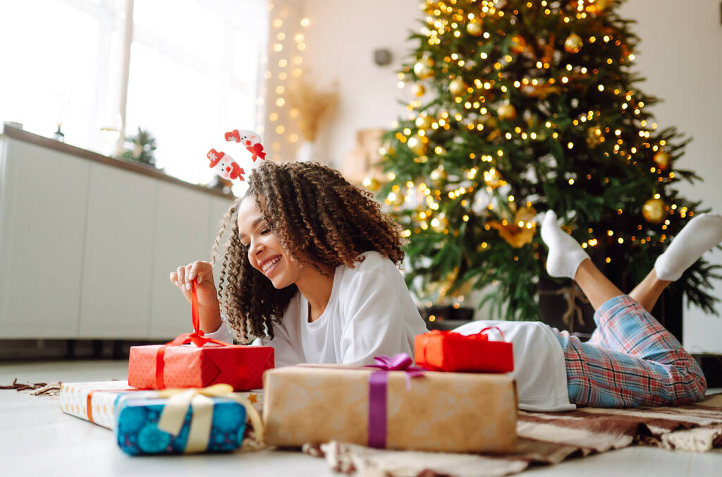 Mujer afroamericana en pijama y sombrero de santa claus con gran caja de regalo posando cerca del árbol de Navidad. Vacaciones de invierno, vacaciones, relax y concepto de estilo de vida. - Foto, Imagen