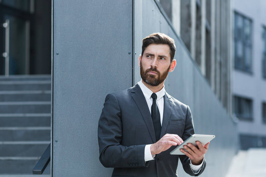 стильный бородатый бизнесмен в формальном деловом костюме стоя с планшетом в руках на заднем плане современного офисного здания снаружи. Человек, использующий смартфон или мобильный телефон на улице города - Фото, изображение