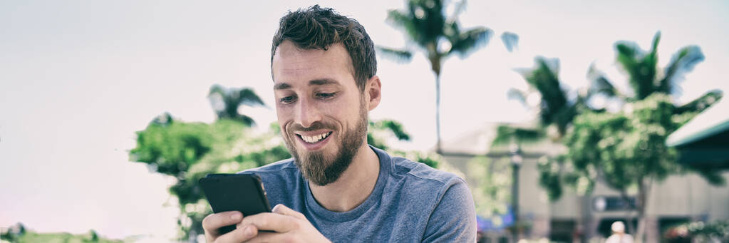 Smartphone man using mobile phone texting sms text message outside in summer background. Young people lifestyle banner panorama. Happy casual guy smiling holding cellphone using app for social media. - Photo, Image