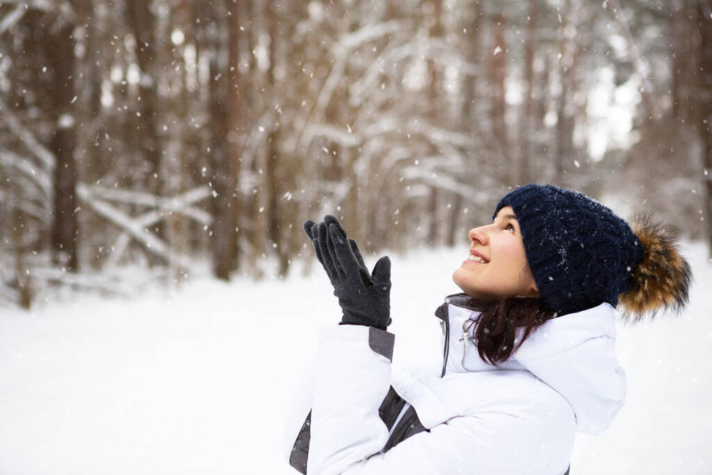 Een vrouw met een warme gebreide hoed over haar ogen getrokken glimlacht en geniet van de sneeuw, koud weer. Outdoor activiteiten, seizoen, winter, sneeuw smelt. Bedekt met vorst, mooie glimlach. Close-up portret - Foto, afbeelding