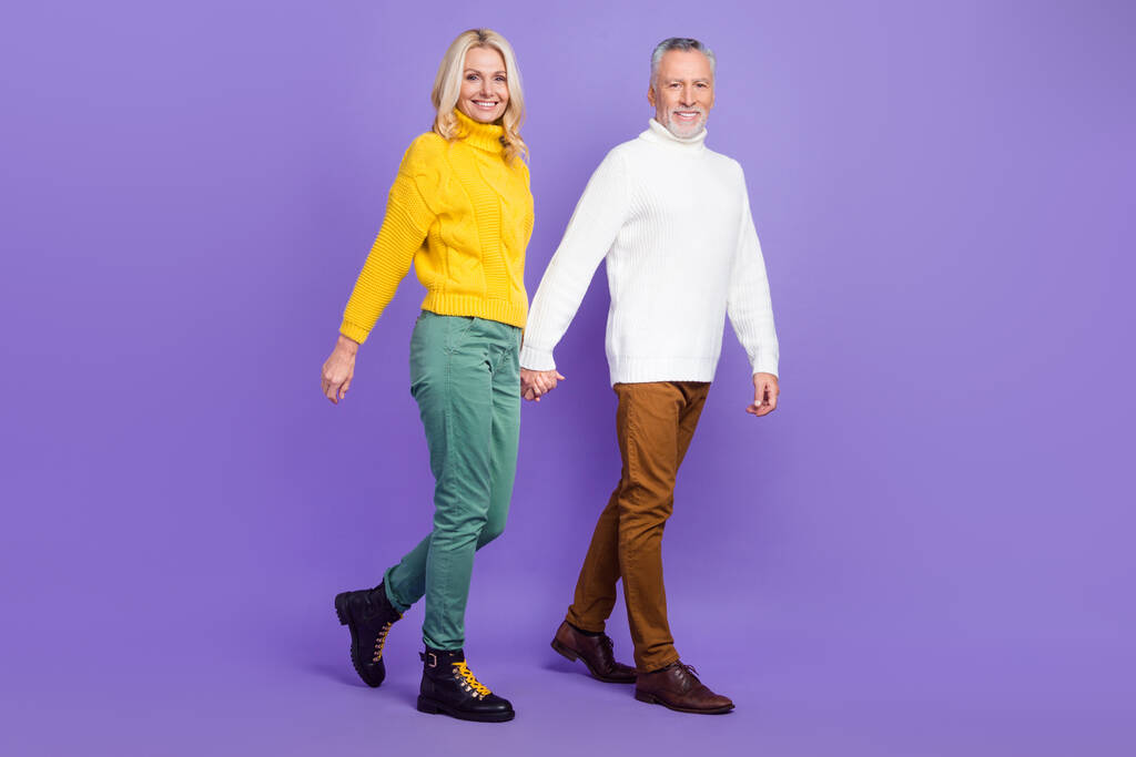 Фото очаровательной взволнованной зрелой жены муж носить вязаные пуловеры улыбаясь ходьба держась руки изолированы фиолетовый цвет фона - Фото, изображение