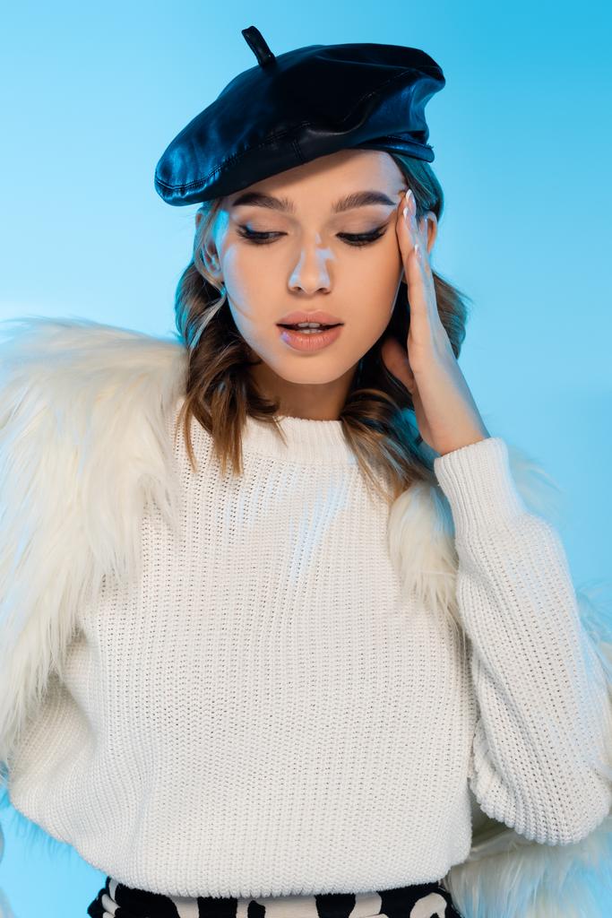 young woman in beret, faux fur jacket and white sweater posing isolated on blue - Photo, Image