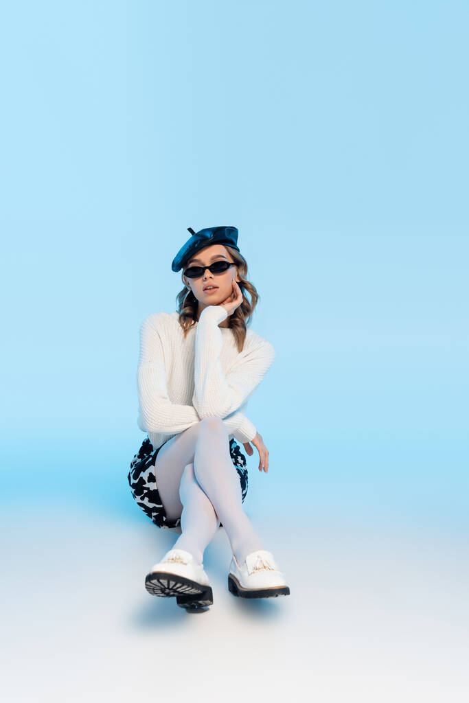 trendi fiatal nő svájcisapkában, napszemüveg és szoknya tehénnyomattal pózol a kék - Fotó, kép