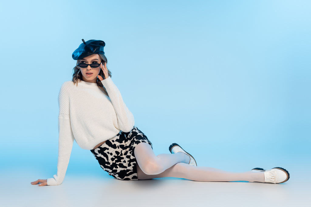 stylová mladá žena v baretu, slunečních brýlích a sukni s kravským potiskem pózující na modré - Fotografie, Obrázek