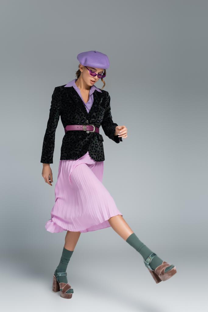 pełna długość stylowa młoda kobieta w berecie, fioletowa spódnica i marynarka z nadrukiem zwierzęcia chodzenie na szarym  - Zdjęcie, obraz