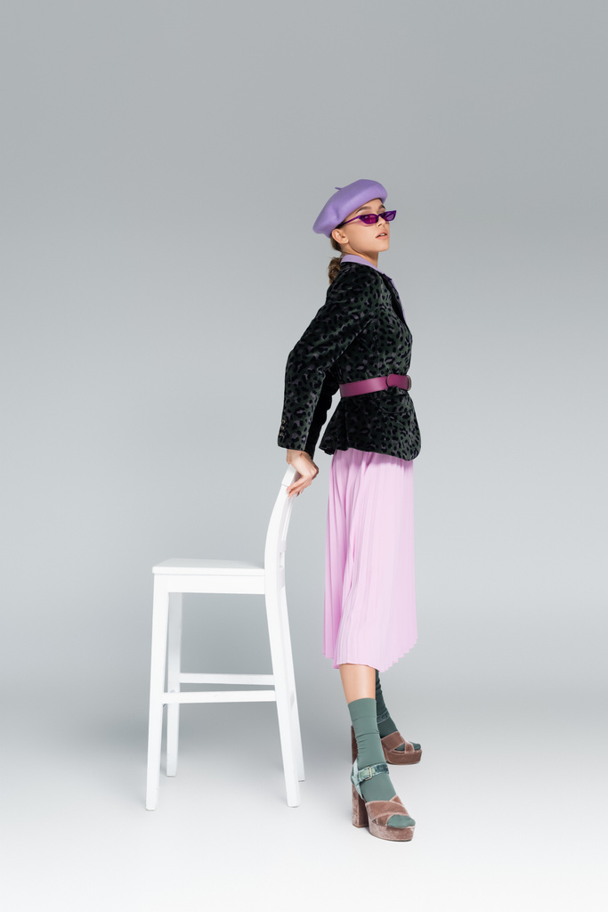 полная длина модной женщины в фиолетовом берете, солнцезащитных очках и пиджаке с отпечатками животных, стоящих рядом со стулом на сером  - Фото, изображение