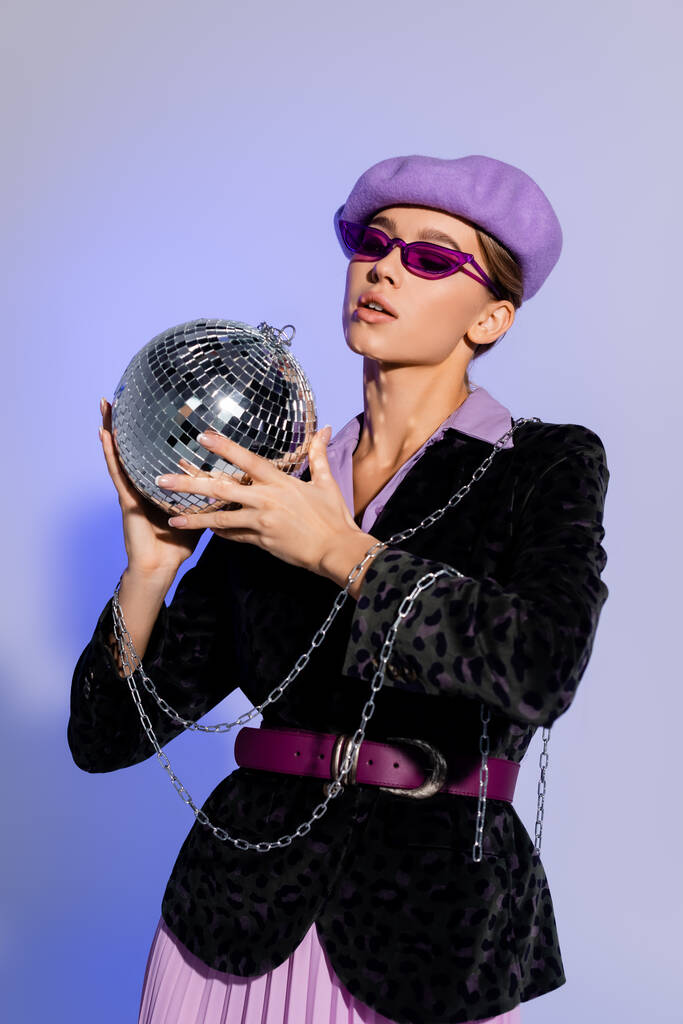 модна жінка в береті, сонцезахисних окулярах і блістері з тваринним принтом, що тримає диско-кулю на фіолетовому
  - Фото, зображення