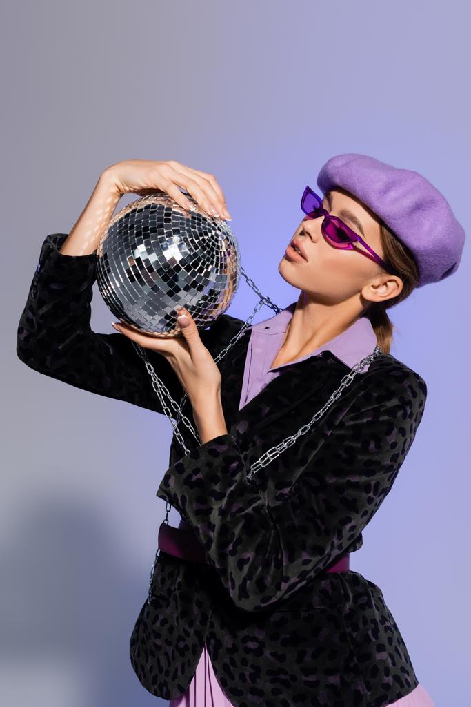 стильная женщина в берете, солнцезащитные очки и пиджак с изображением животных глядя на диско мяч на фиолетовый  - Фото, изображение