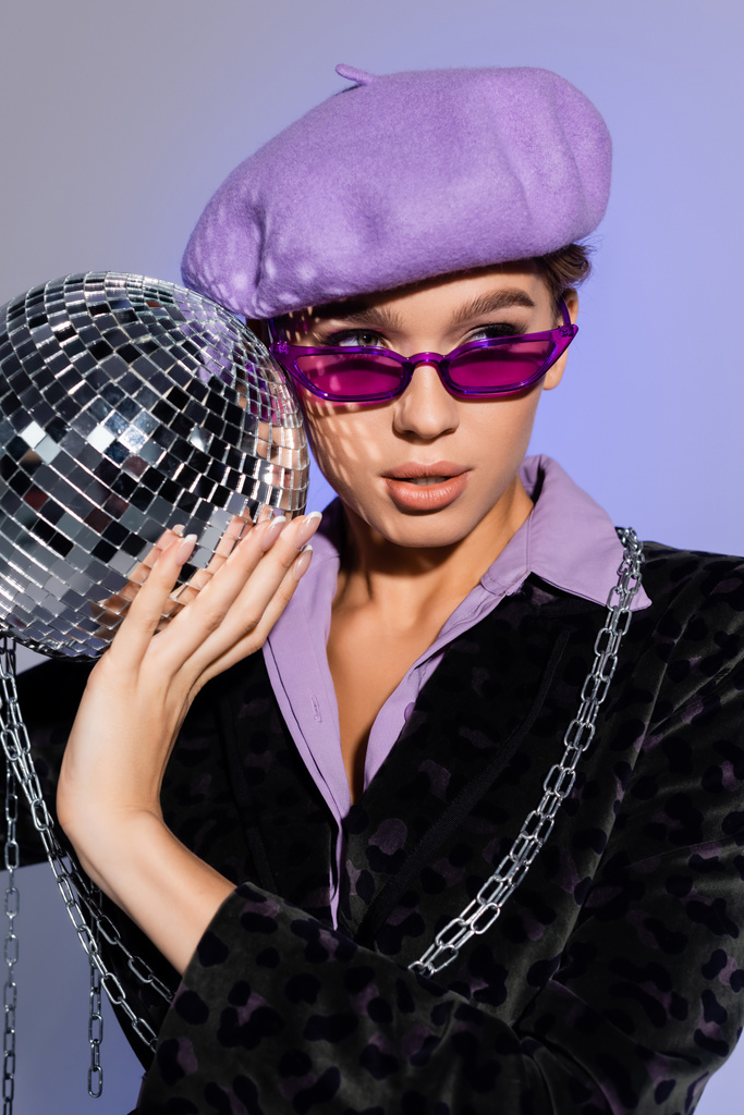 jovem mulher elegante em boina, óculos de sol e blazer com animal imprimir segurando bola de discoteca brilhante em roxo  - Foto, Imagem