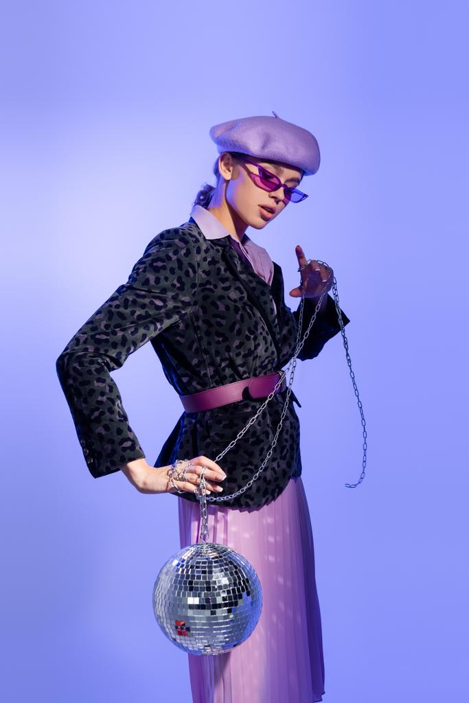 stylische Frau in Baskenmütze, Sonnenbrille und Blazer mit Animal Print hält Discokugel an Kette auf violett - Foto, Bild