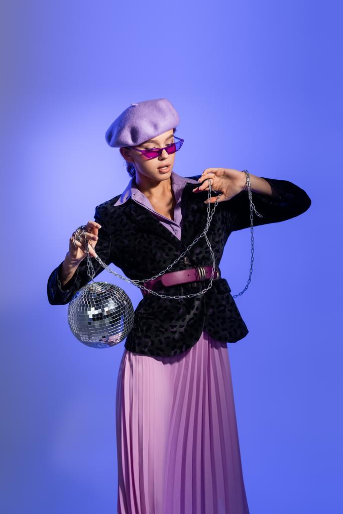 femme élégante en béret violet, lunettes de soleil et blazer avec imprimé animal tenant boule disco sur chaîne sur violet - Photo, image