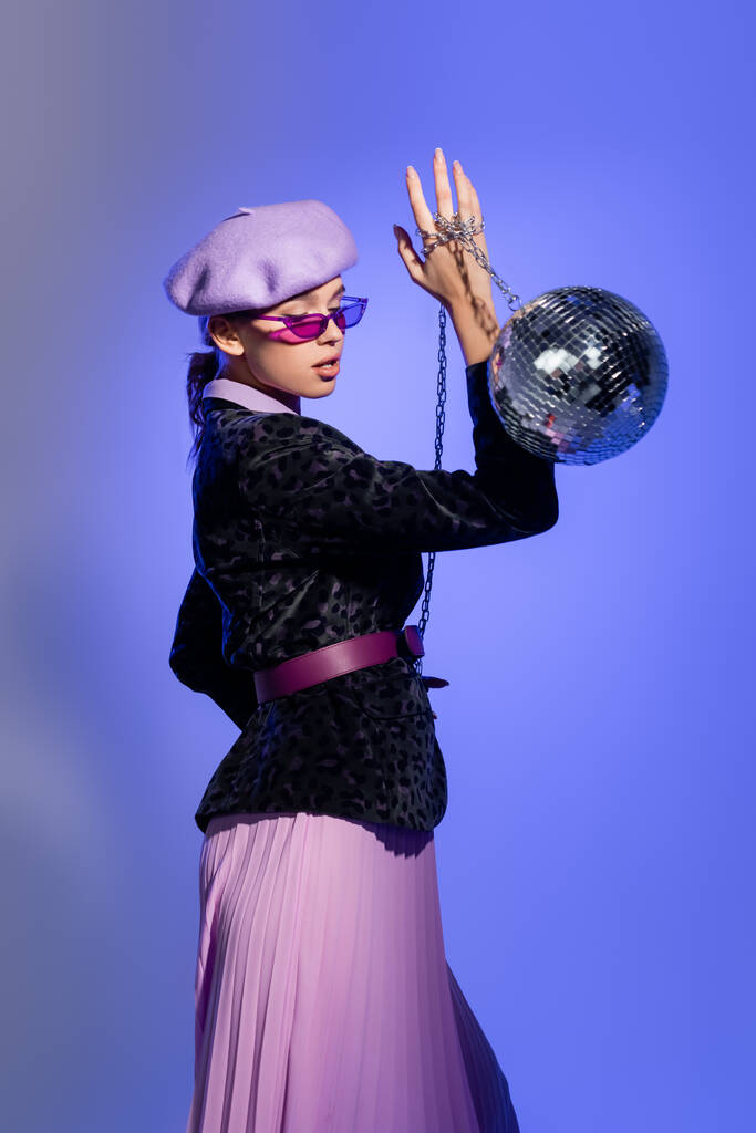 κομψή γυναίκα σε μωβ μπερέ και σακάκι με animal print κρατώντας disco μπάλα στην αλυσίδα σε βιολέτα - Φωτογραφία, εικόνα