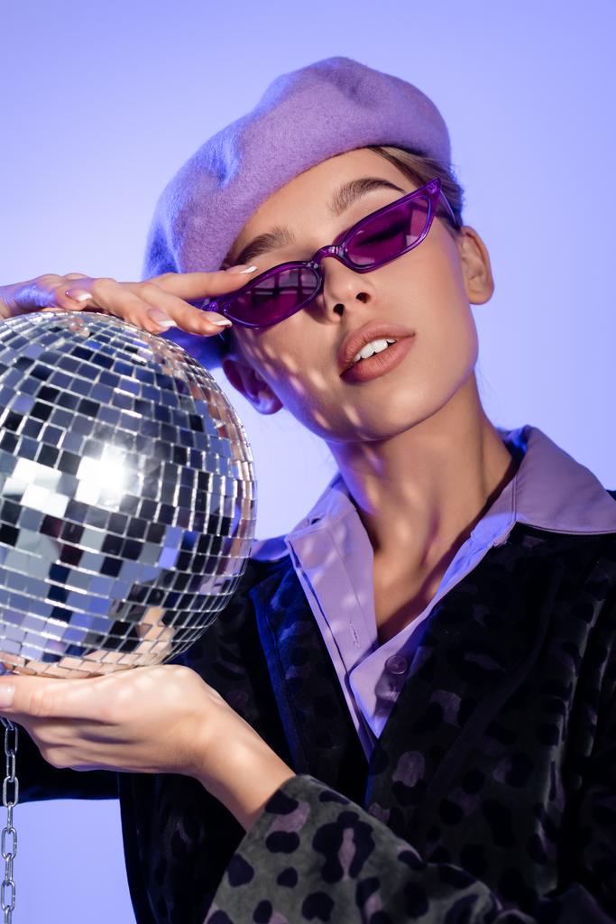 μοντέρνα γυναίκα σε μπερέ και γυαλιά ηλίου κρατώντας disco μπάλα στην αλυσίδα σε βιολέτα - Φωτογραφία, εικόνα