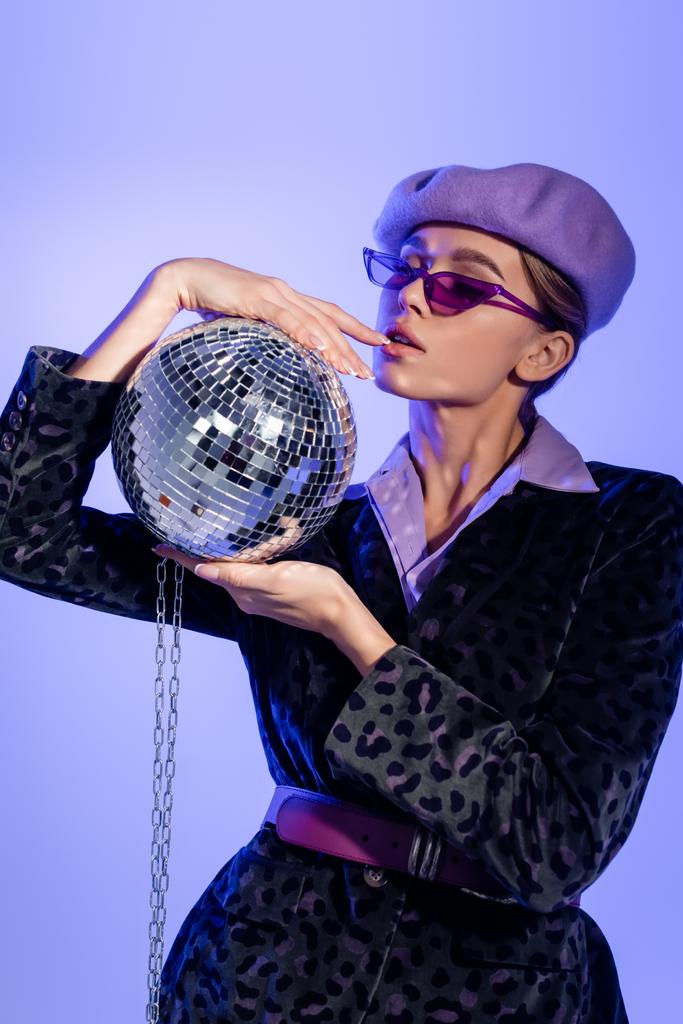 young woman in purple beret, sunglasses and blazer with animal print holding disco ball on chain on violet - Photo, Image