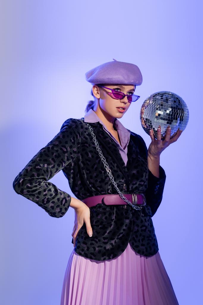 giovane donna in basco viola e blazer con stampa animalier che tiene la palla da discoteca mentre posa con mano sull'anca su viola - Foto, immagini