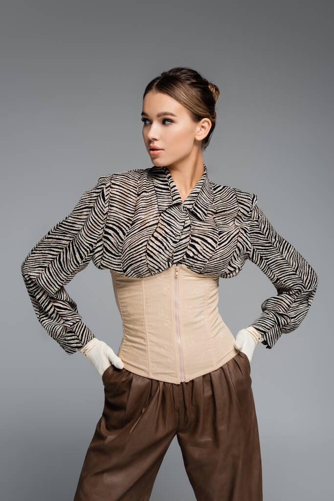 trendy woman in blouse with animal print, gloves and corset posing with hands in pockets isolated on grey - Photo, Image