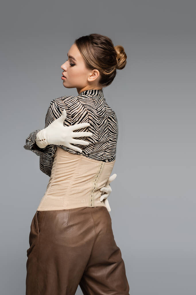 μοντέρνα γυναίκα σε μπλούζα με animal print, γάντια και κορσέ αγκαλιάζει τον εαυτό της απομονωμένη σε γκρι - Φωτογραφία, εικόνα