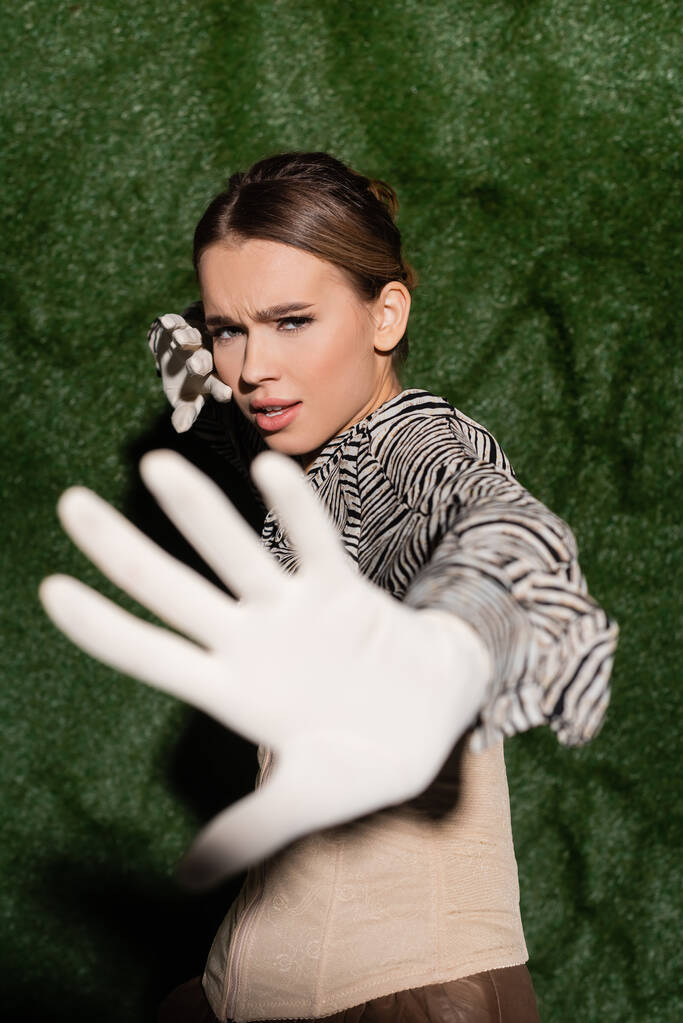 młody modny model w bluzce z nadrukiem zebry i rękawiczkami niepokazującymi gestu w pobliżu trawiastego tła  - Zdjęcie, obraz