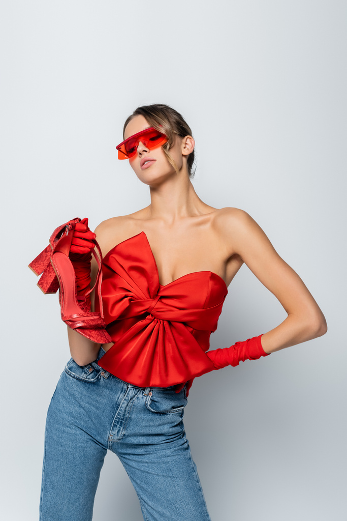 赤のブラウスとサングラスと手袋を手でポーズしながら靴を保持するスタイリッシュなモデルグレーに隔離されたヒップ - 写真・画像