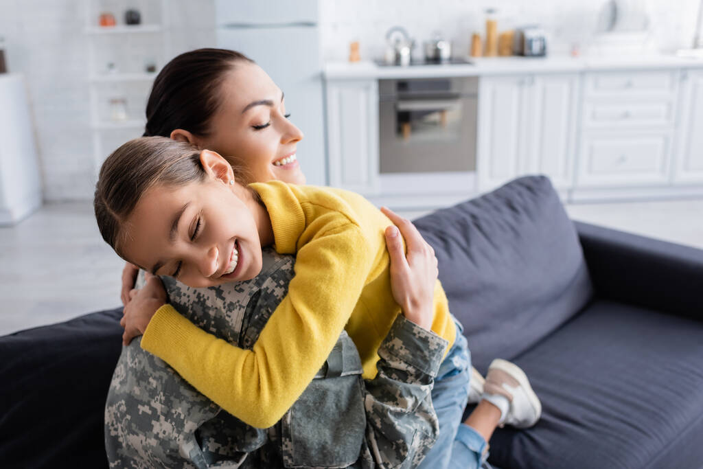 Усміхнена жінка в військовій формі закриває очі, обіймаючи доньку вдома
  - Фото, зображення