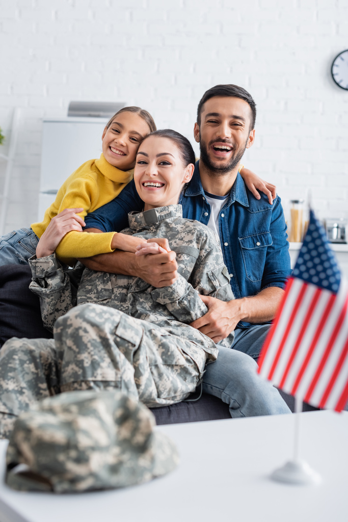 Felice famiglia abbracciando mamma in uniforme militare e guardando la fotocamera vicino bandiera americana a casa  - Foto, immagini