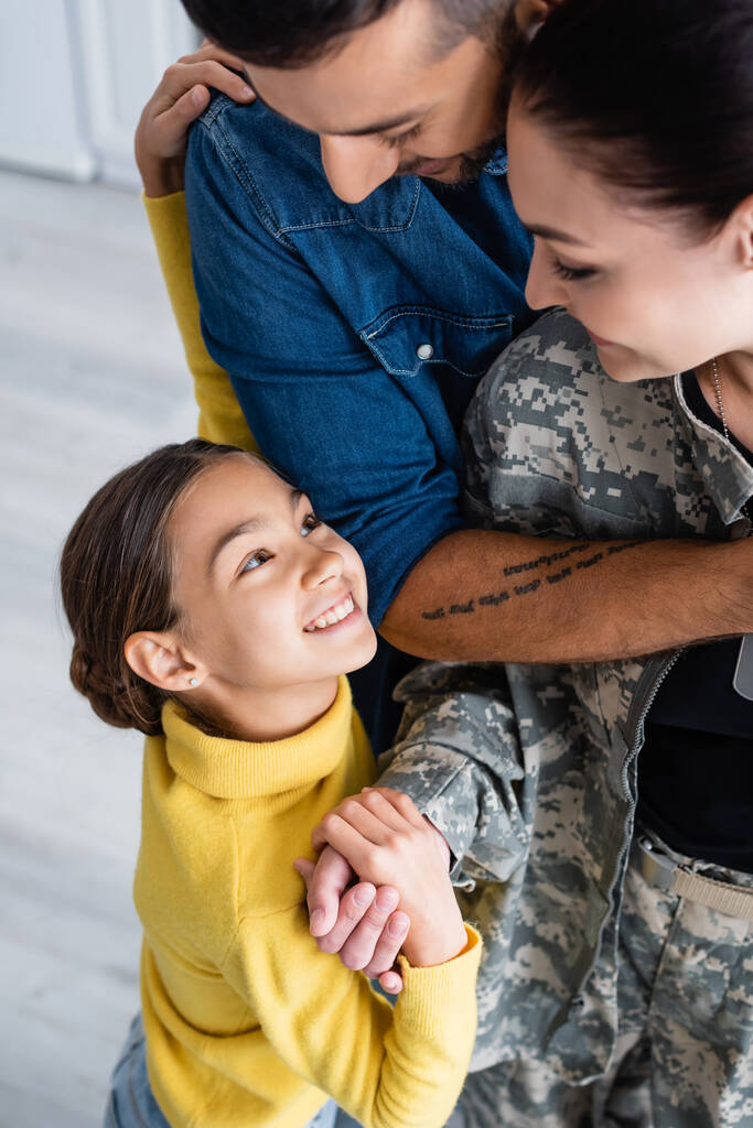 Positive woman in camouflage uniform holding hand of preteen daughter near husband at home  - Photo, Image