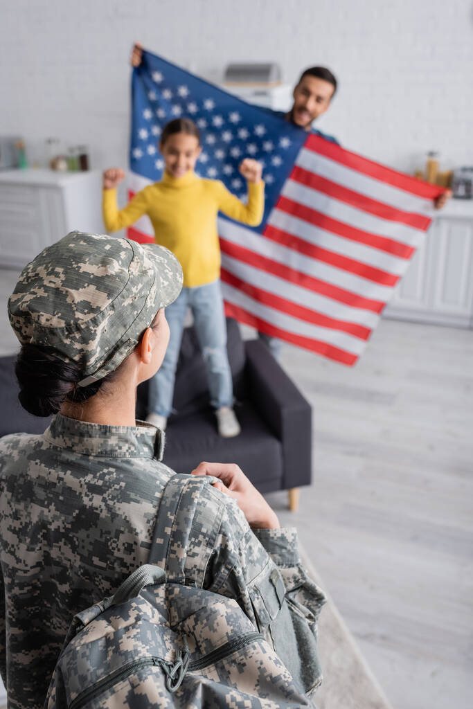 Askeri üniformalı bir kadın. Sırt çantası yanında bulanık bir aile. Mutfakta Amerikan bayrağı taşıyor.  - Fotoğraf, Görsel