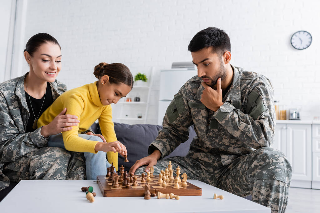 Padre pensieroso in uniforme mimetica giocare a scacchi vicino sorridente mamma e bambino a casa  - Foto, immagini