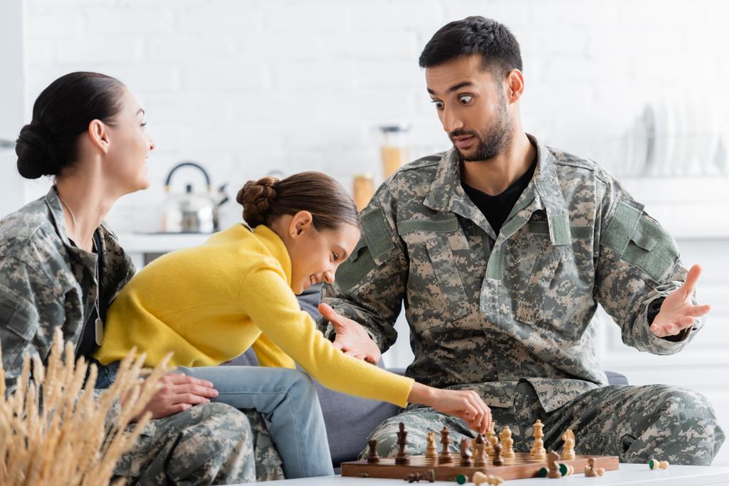 Hombre sorprendido en uniforme de camuflaje jugando ajedrez con la familia en casa  - Foto, imagen