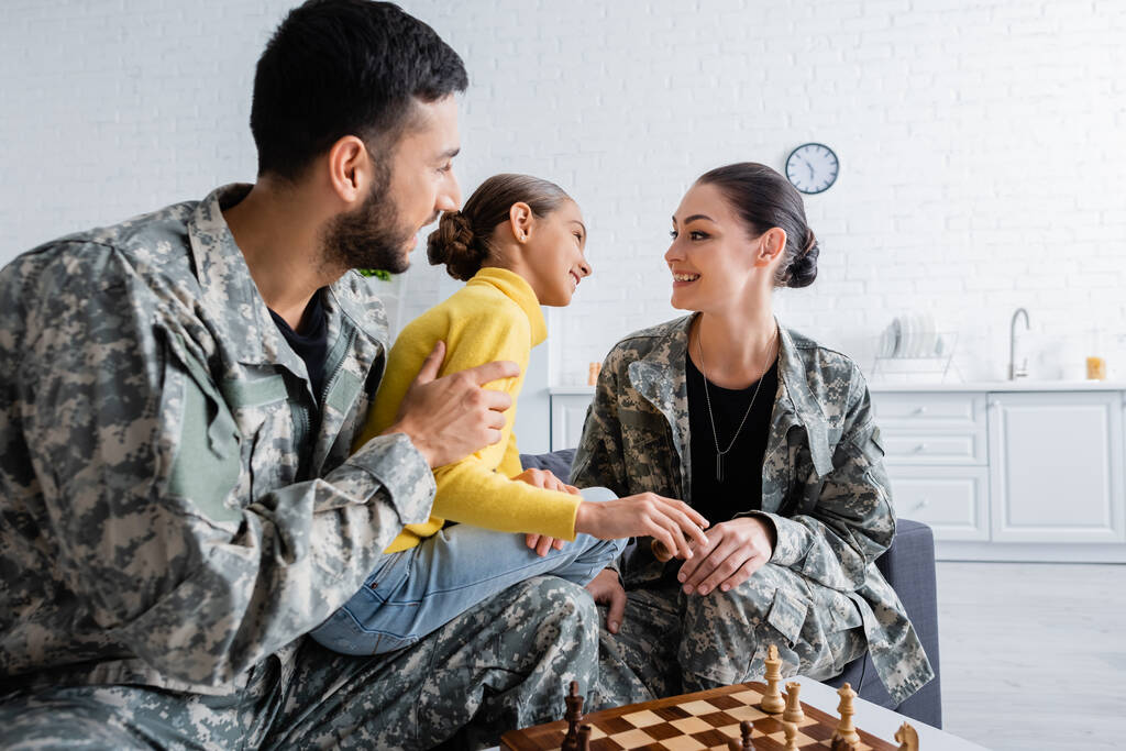 Positive Eltern in Militäruniform sitzen neben Tochter und Schach zu Hause  - Foto, Bild