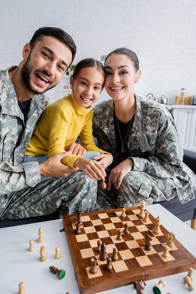 Улыбающиеся родители в военной форме смотрят в камеру рядом с ребенком с фигурой шахматиста на диване дома  - Фото, изображение