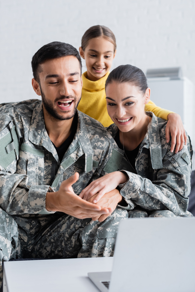 Χαμογελώντας γονείς με στρατιωτική στολή κρατώντας τα χέρια κοντά στο φορητό υπολογιστή και την κόρη στο σπίτι  - Φωτογραφία, εικόνα