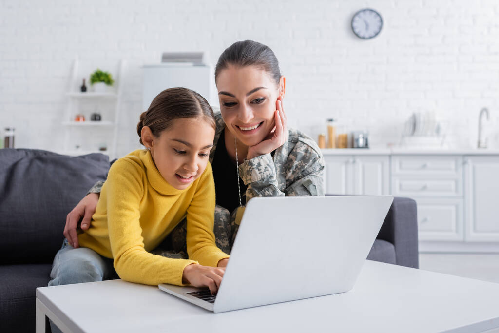 Усміхнена дівчина використовує ноутбук біля матері у військовій формі вдома
  - Фото, зображення