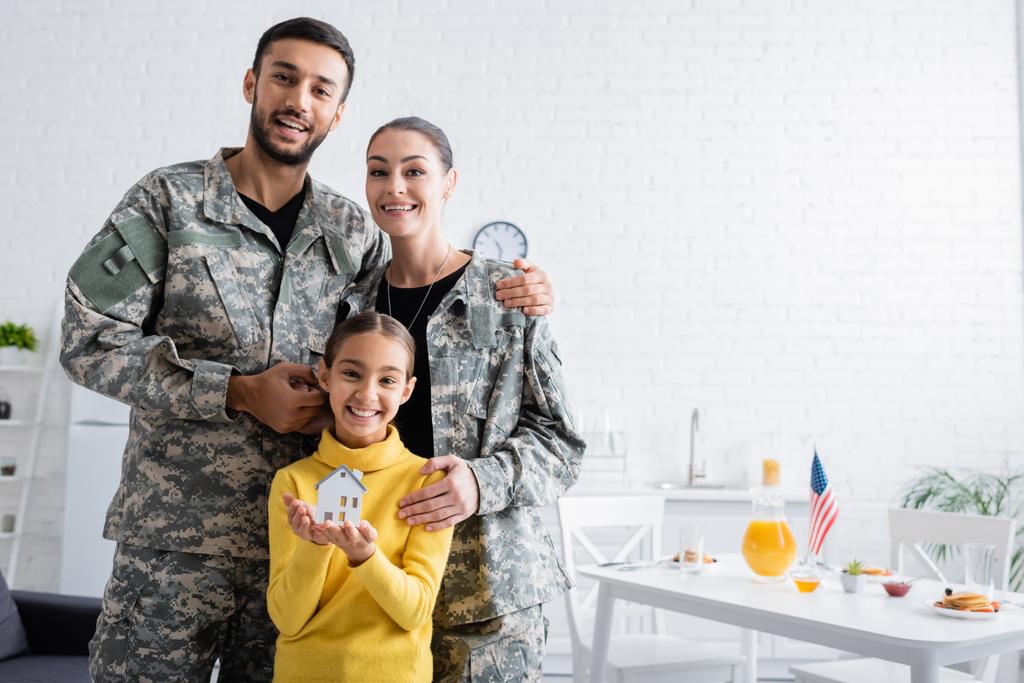 Parents souriants en uniforme militaire debout près de la fille avec modèle de maison dans la cuisine  - Photo, image