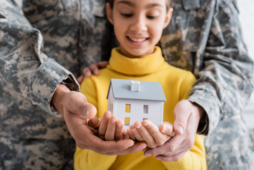 Modell eines Hauses auf Händen eines verschwommenen Kindes in der Nähe von Eltern in Militäruniform zu Hause  - Foto, Bild