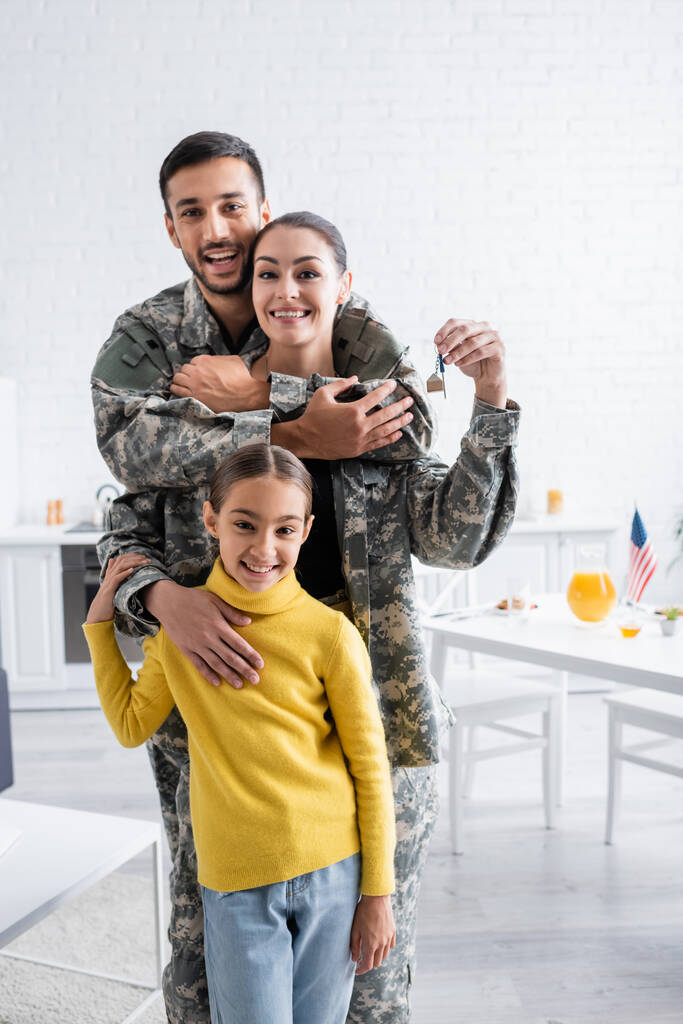 Sonriente hombre en uniforme militar abrazando esposa con llave cerca de niño en casa  - Foto, Imagen