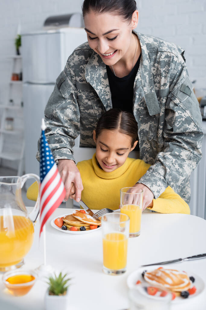 Χαμογελαστή γυναίκα με στολή καμουφλάζ κόβει τηγανίτες κοντά στην κόρη και την αμερικανική σημαία στην κουζίνα   - Φωτογραφία, εικόνα