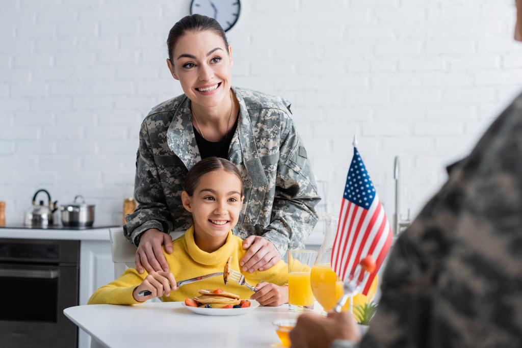 Usmívající se matka v maskovací uniformě stojící poblíž dcery a americké vlajky při snídani v kuchyni  - Fotografie, Obrázek