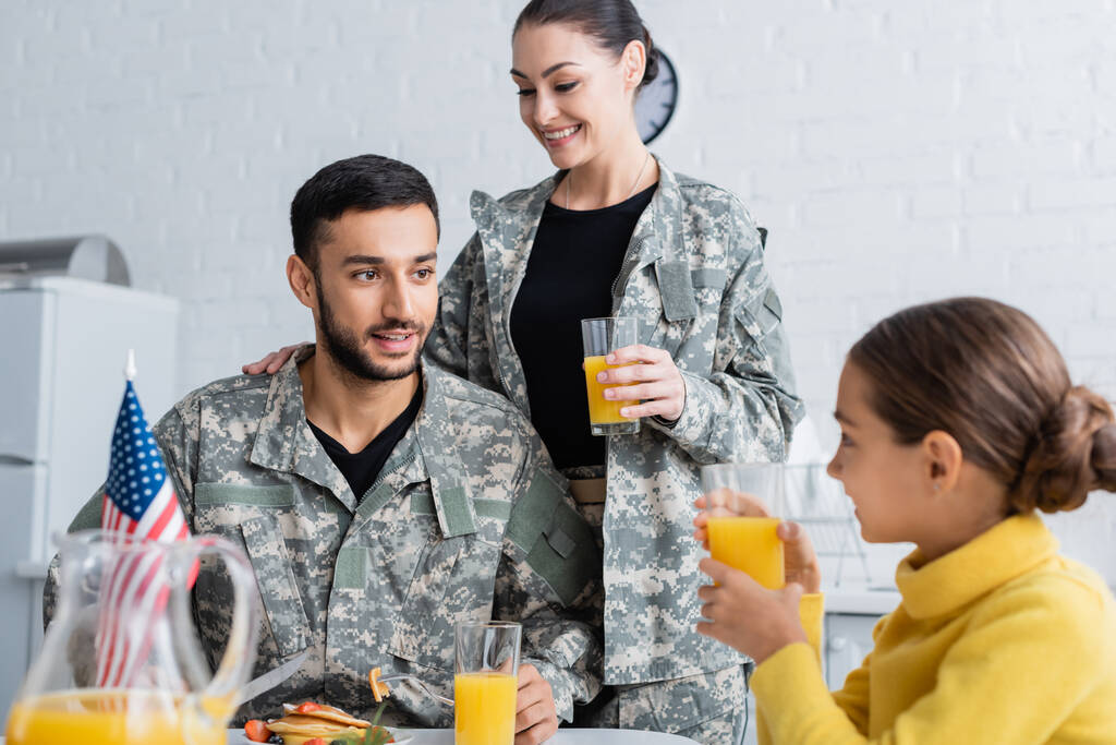 Des parents souriants en uniforme militaire regardant leur fille près du drapeau américain pendant le petit déjeuner à la maison  - Photo, image