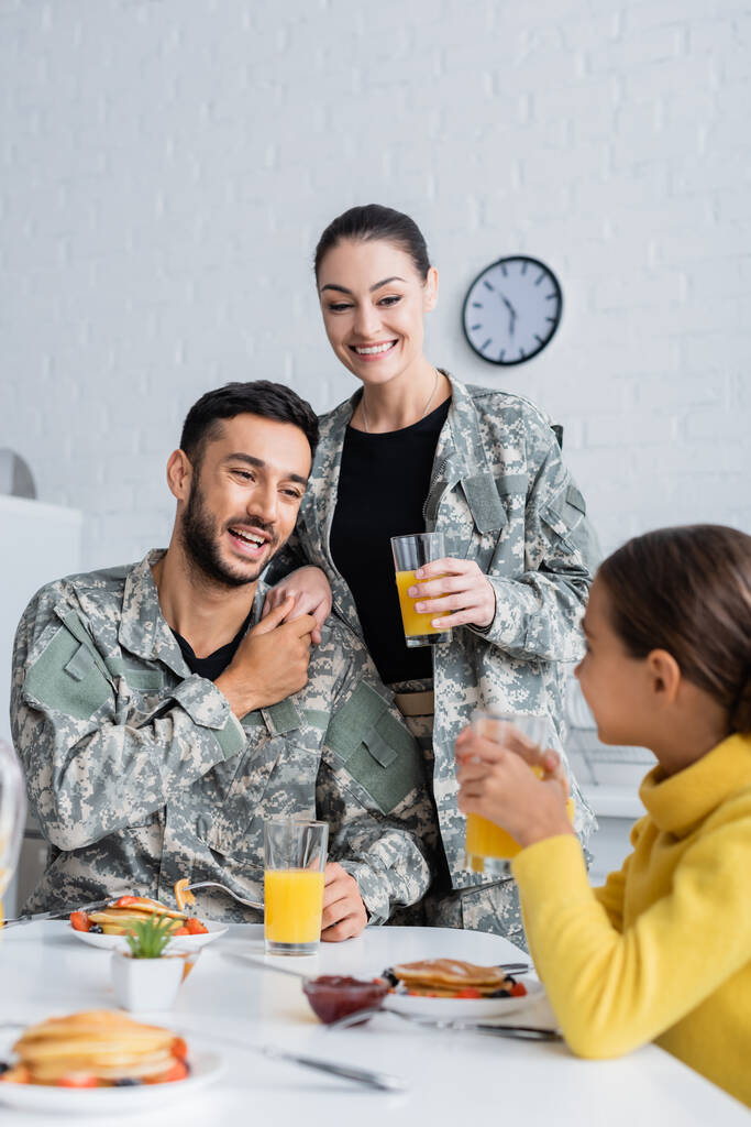 Vidám szülők katonai egyenruhában, egymás kezét fogva a lányuk mellett reggeliznek otthon.  - Fotó, kép