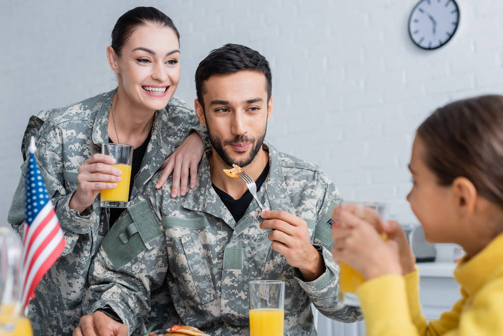 pais sorridentes em uniforme militar olhando para a criança perto da bandeira americana durante o café da manhã em casa  - Foto, Imagem