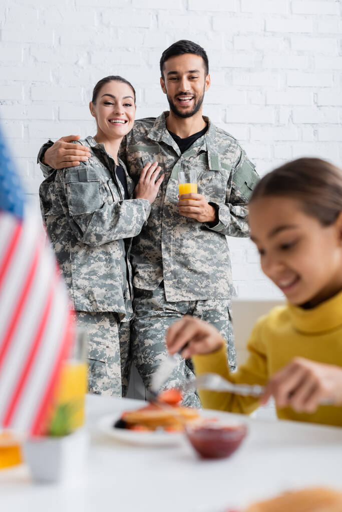 Alegres padres en uniforme de camuflaje mirando a su hija desayunando cerca de la borrosa bandera americana en casa  - Foto, imagen