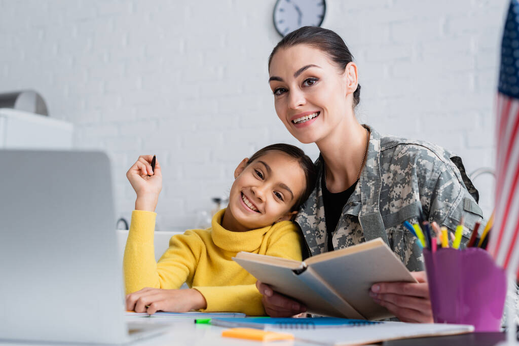 Femme souriante en uniforme militaire tenant le livre près de l'enfant, ordinateur portable et drapeau américain à la maison  - Photo, image
