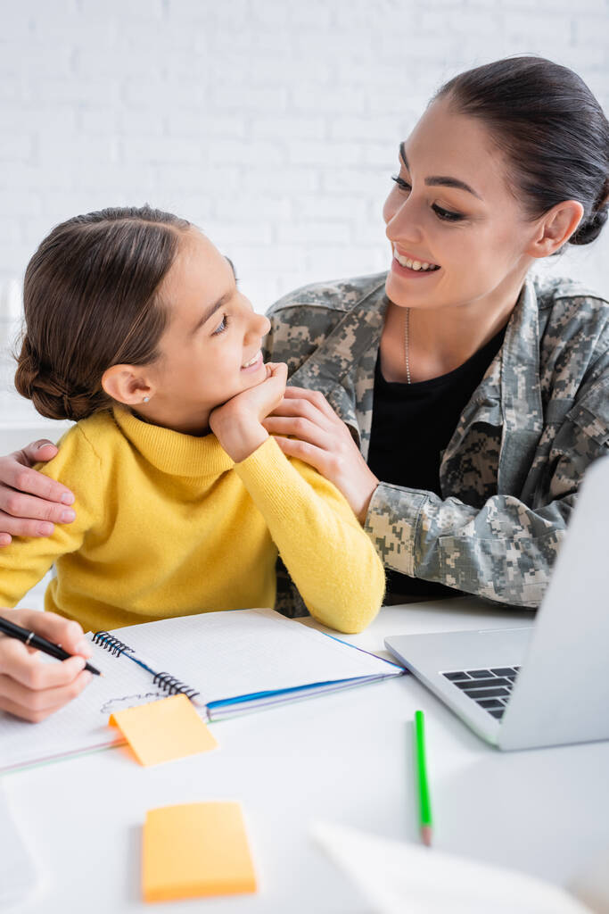 Uśmiechnięta matka w mundurze wojskowym patrząca na dziecko podczas lekcji w pobliżu laptopa w domu  - Zdjęcie, obraz