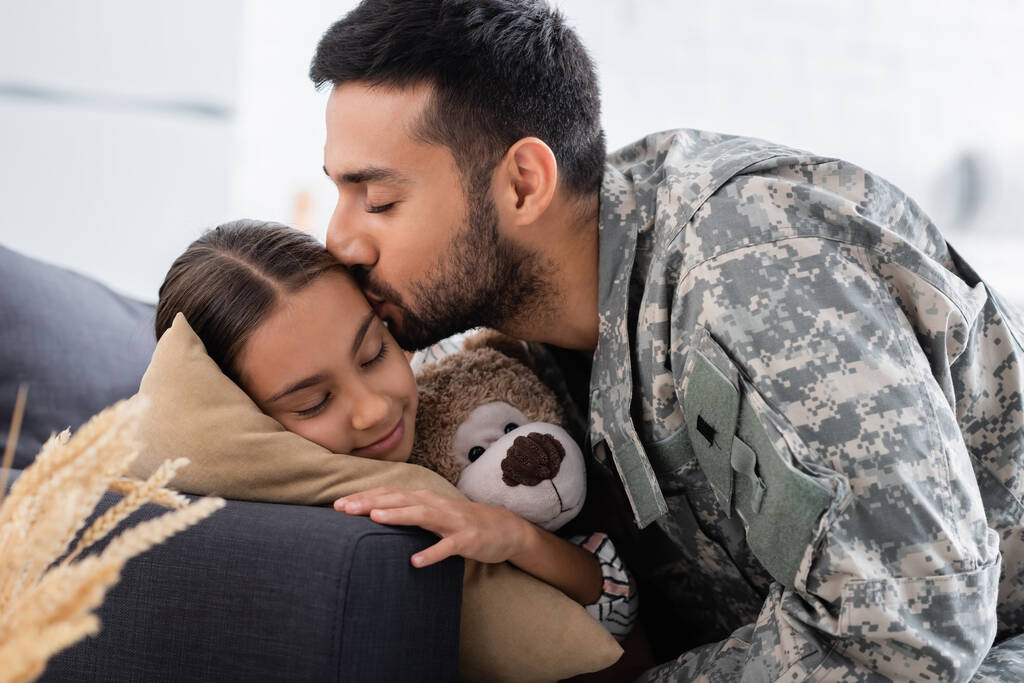Man in military uniform kissing child with teddy bear on couch  - Photo, Image