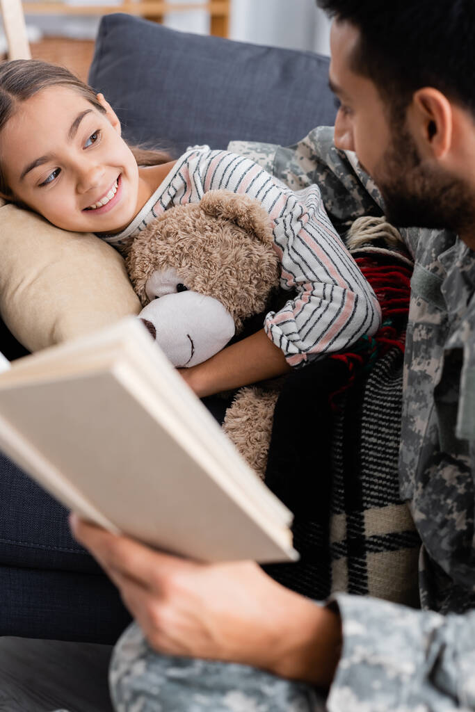Boldog gyermek plüssmackóval nézi az elmosódott apát katonai egyenruhában, aki otthon tartja a könyvet.  - Fotó, kép