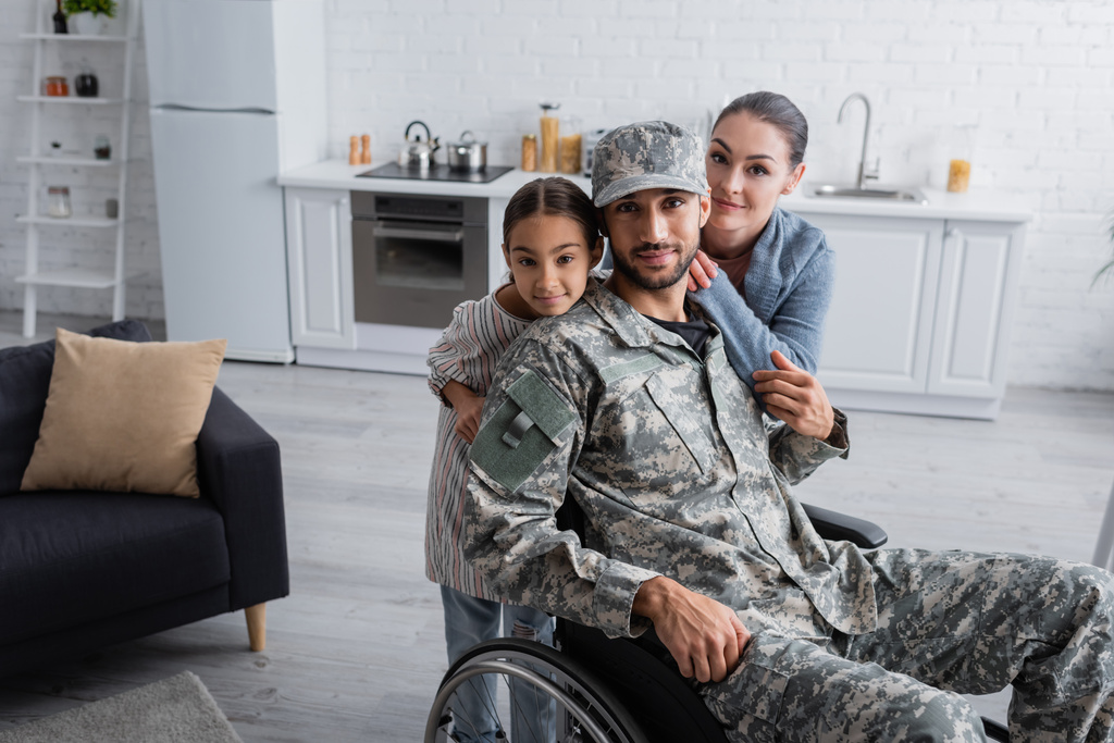 Mężczyzna w mundurze wojskowym i wózku inwalidzkim patrząc na kamerę w pobliżu rodziny w domu  - Zdjęcie, obraz