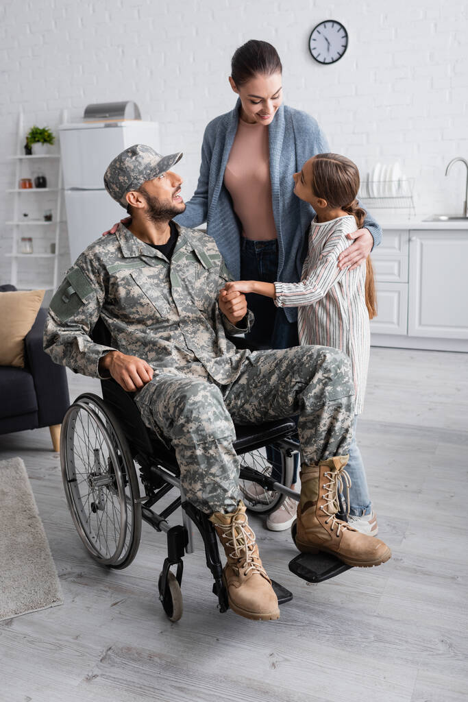 Wesoła kobieta i dziecko stojące obok ojca w mundurze wojskowym i wózku inwalidzkim w domu  - Zdjęcie, obraz