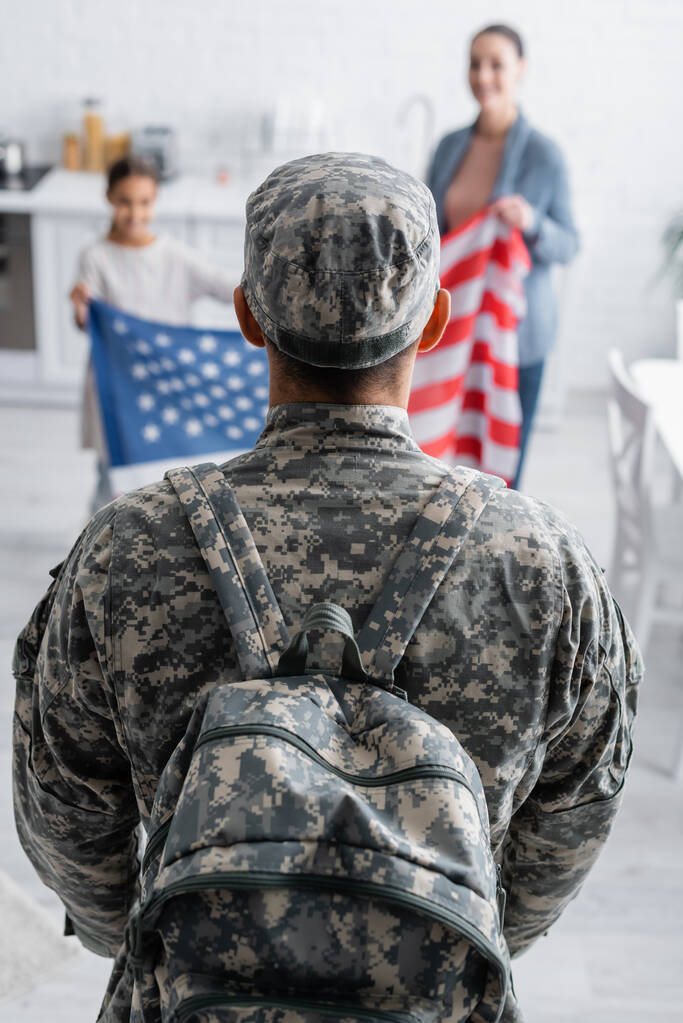 Hombre en uniforme de camuflaje con mochila cerca de la familia borrosa con bandera americana en casa  - Foto, Imagen