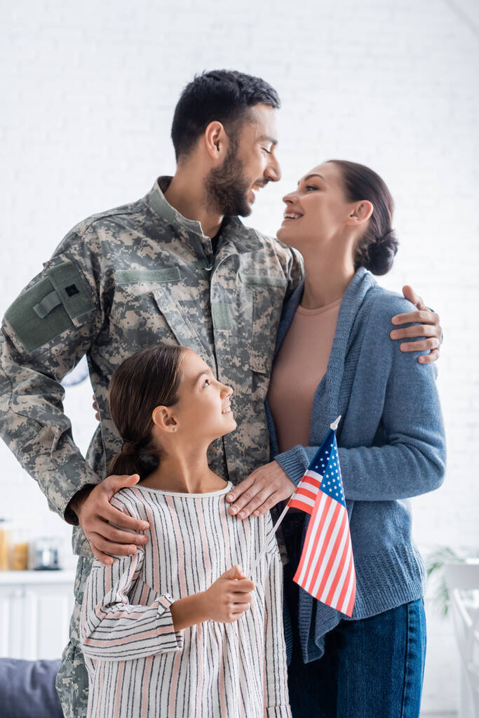 Pozytywna kobieta patrząc na męża w mundurze kamuflażu w pobliżu córki z amerykańską flagą w domu  - Zdjęcie, obraz